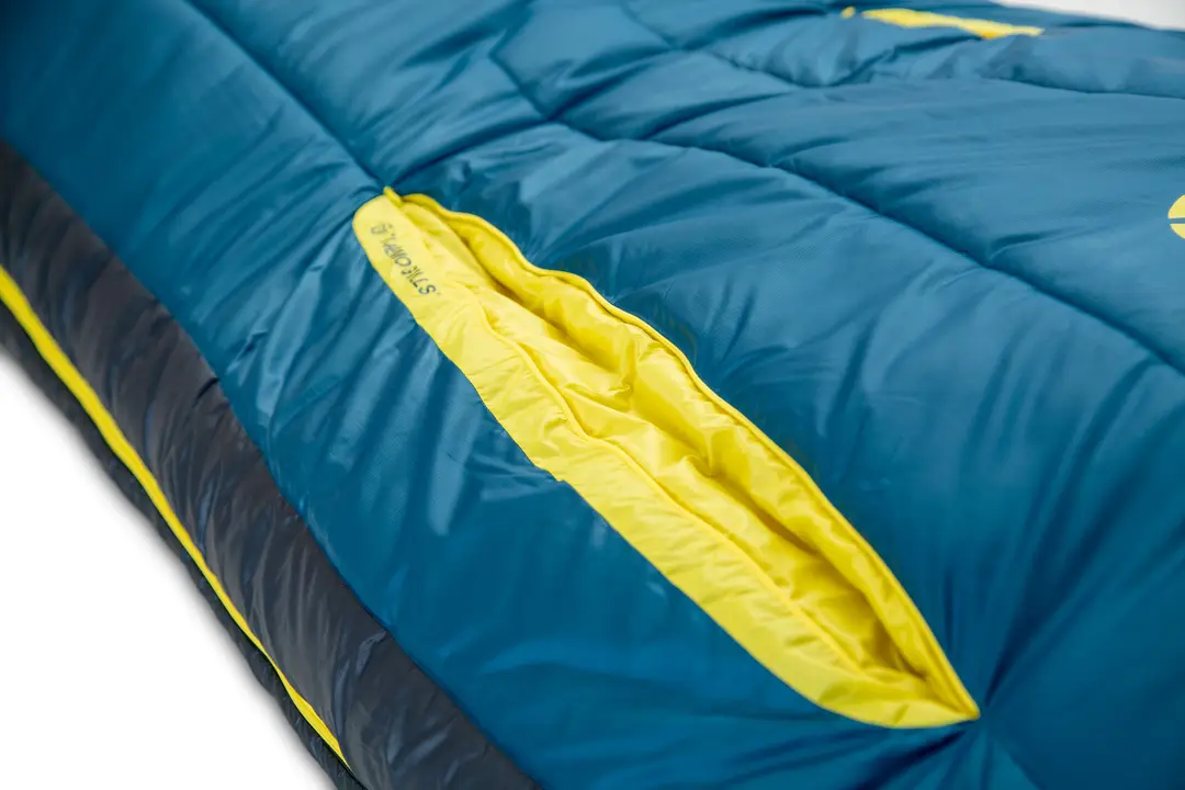 Gros plan d'un sac de couchage bleu aux touches de jaune, avec une fermeture éclair visible sur une bande jaune sur le côté. Le tissu semble matelassé et matelassé pour une meilleure isolation.