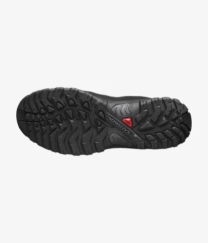 L'image montre la semelle d'une chaussure de randonnée noire conçue avec des crampons profonds et robustes pour une meilleure traction. La semelle présente un petit triangle rouge et le nom de la marque « Salomon » gravé en lettres en relief.