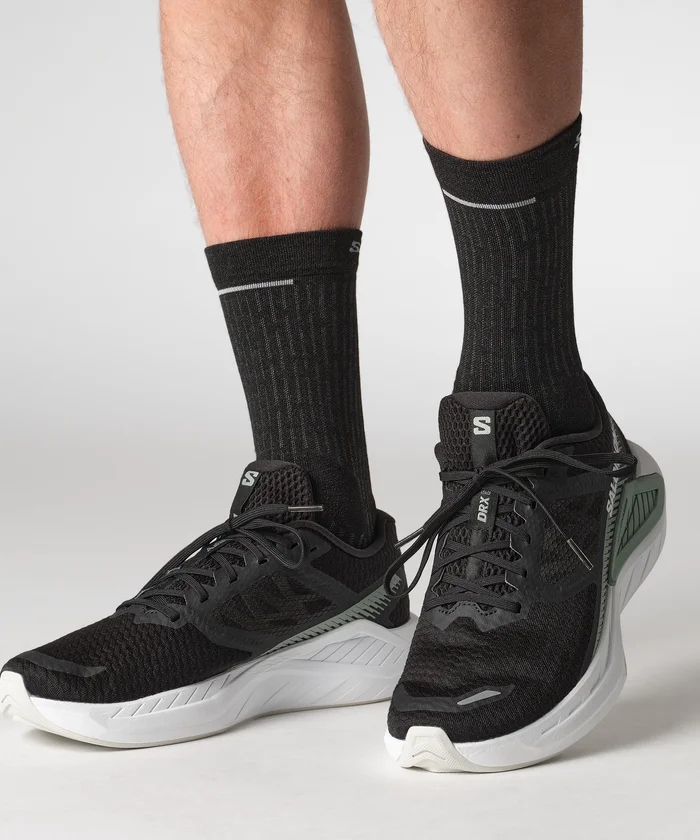 Gros plan d'une personne portant des chaussures de sport noires à semelles blanches et des chaussettes noires. Les chaussures présentent un design texturé avec des accents verts et les chaussettes ont de petits motifs subtils. L'arrière-plan est simple et minimaliste.