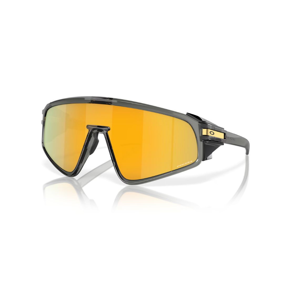 La LUNETTE DE SOLEIL OAKLEY LATCH PANEL se caractérise par une monture fumée grise et de grands verres angulaires PRIZM 24K miroités, présentés sur un fond blanc avec un reflet subtil en dessous.