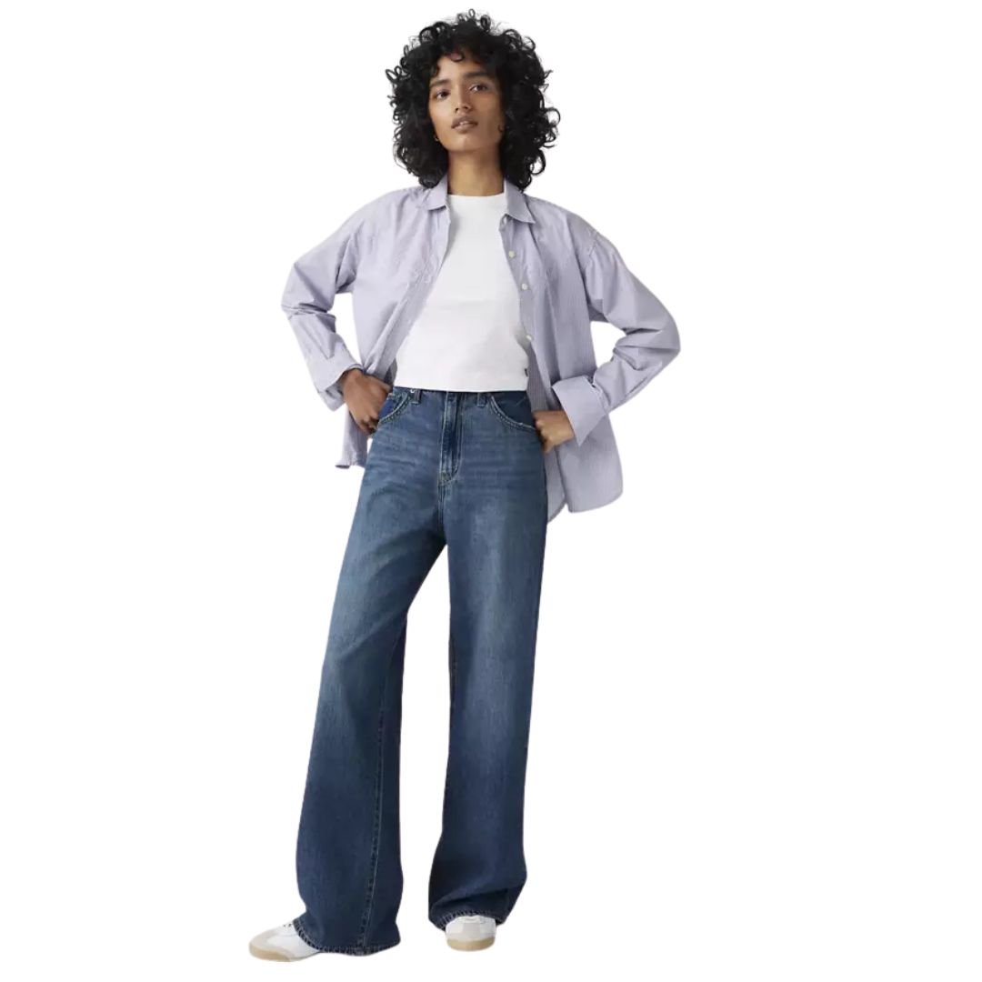 Une personne aux cheveux bouclés se tient debout, les mains sur les hanches, vêtue d'un jean bleu à jambe large CINCH BAGGY, d'une chemise à boutons violet clair sur un t-shirt blanc et de chaussures blanches sur un fond blanc uni.