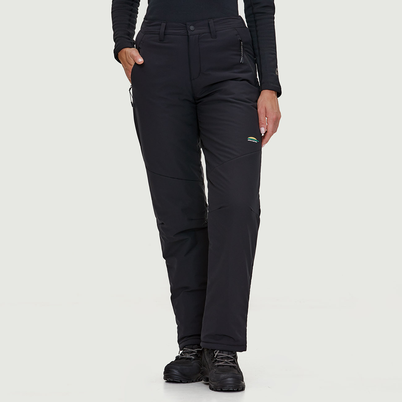 Une personne portant un pantalon de neige noir et des bottes noires se tient debout sur un fond uni. Le pantalon a des poches zippées et un petit logo sur le haut de la cuisse. Les bottes sont à lacets et robustes.