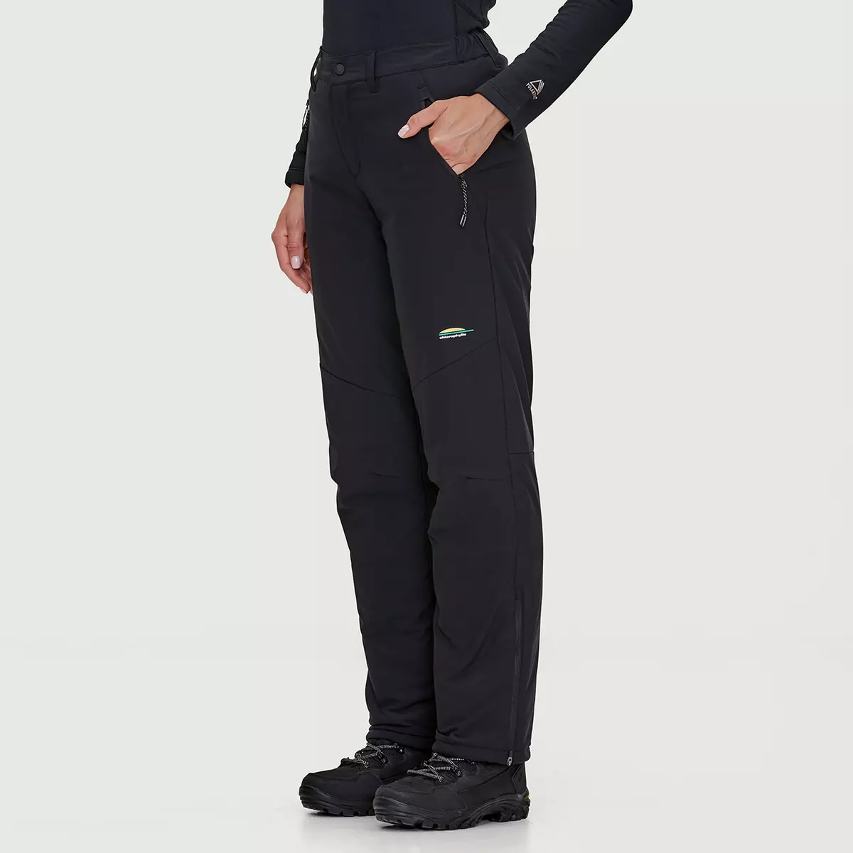 Une personne porte un pantalon d'extérieur noir et une chemise à manches longues sur fond clair. Le pantalon comporte des fermetures éclair et un petit logo sur la cuisse. La personne porte également des chaussures de randonnée noires.