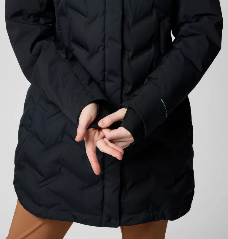 Une personne porte une veste matelassée noire, les mains croisées devant elle. La veste a un col haut et les doigts de la personne sont entrelacés. La partie inférieure de la veste est visible sur un pantalon marron.