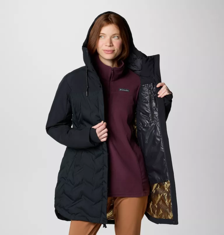Une personne aux cheveux longs porte un manteau d'hiver noir à capuche et laisse apparaître sa doublure intérieure. Elle porte également une chemise marron sous le manteau et un pantalon marron clair. L'arrière-plan est d'une couleur unie, gris clair.