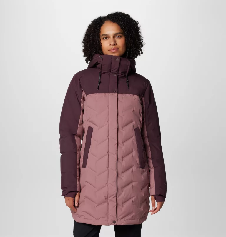Une personne porte un manteau d'hiver à capuche bicolore violet et rose sur un fond uni. Le manteau présente un motif à chevrons et des boutons foncés sur le devant.