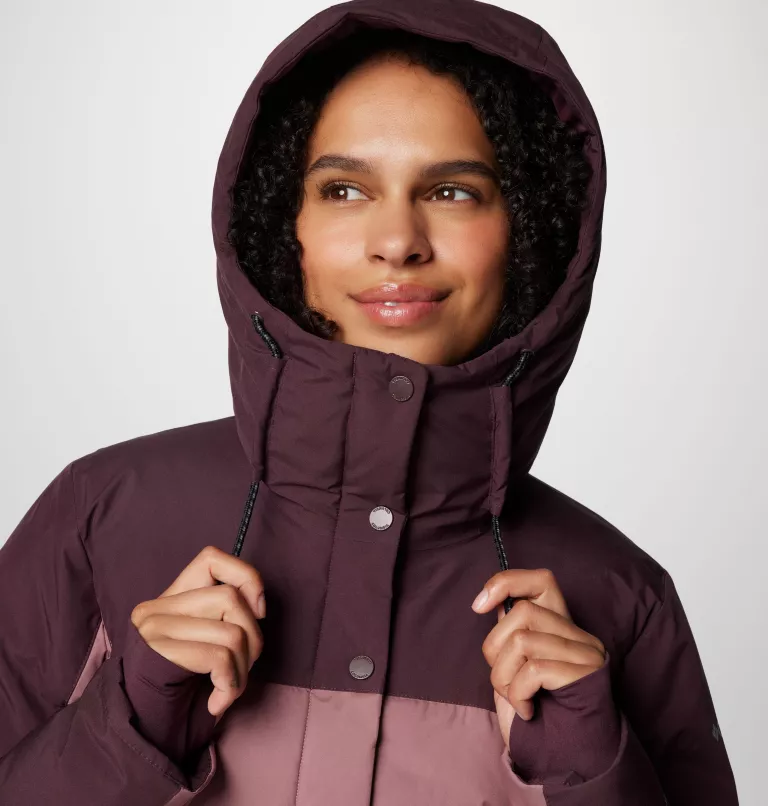 Une personne aux cheveux bouclés porte une veste bicolore, violet foncé et violet clair, avec une capuche relevée. La personne regarde sur le côté avec un léger sourire, tenant le col de la veste. Le fond est uni et de couleur claire.