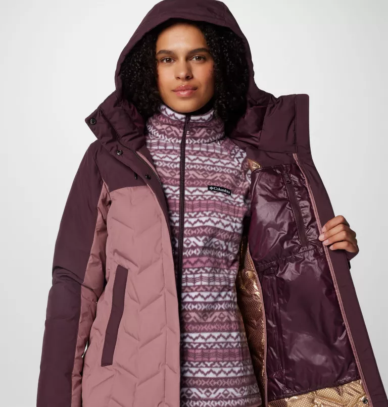 Une personne porte une veste d'hiver bicolore avec une capuche ouverte, révélant la couche intérieure en polaire à motifs. La veste est marron et rose avec un motif géométrique. Le fond est blanc uni.