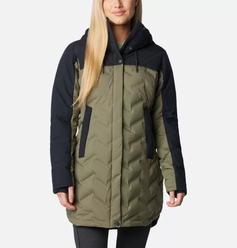 Une personne porte une veste bicolore avec des parties vert olive et noires. La veste a une capuche, des manches noires, des poches zippées et un motif matelassé. La personne a de longs cheveux blonds et se tient debout sur un fond uni.