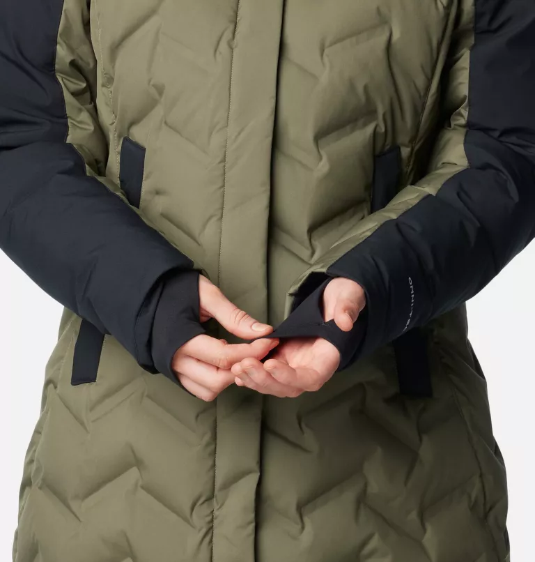 Une personne portant une veste matelassée verte à manches noires est représentée en train d'ajuster les poignets. La veste est boutonnée et la personne utilise ses doigts pour tirer ou ajuster le tissu au niveau du poignet.