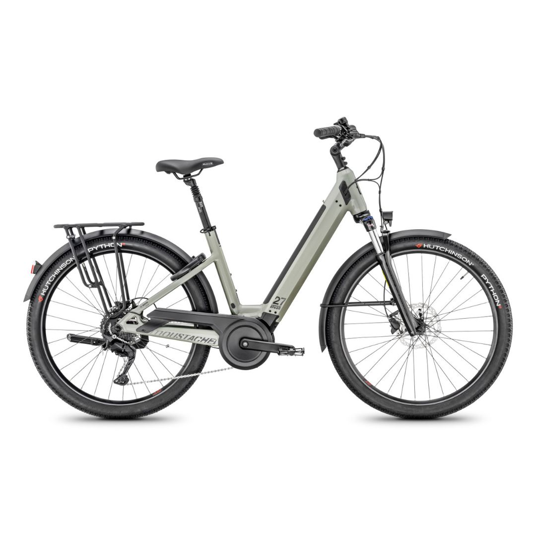 Un vélo électrique gris avec un cadre traversant, une suspension avant, un porte-bagages arrière et des garde-boue noirs, présenté de côté sur fond blanc.