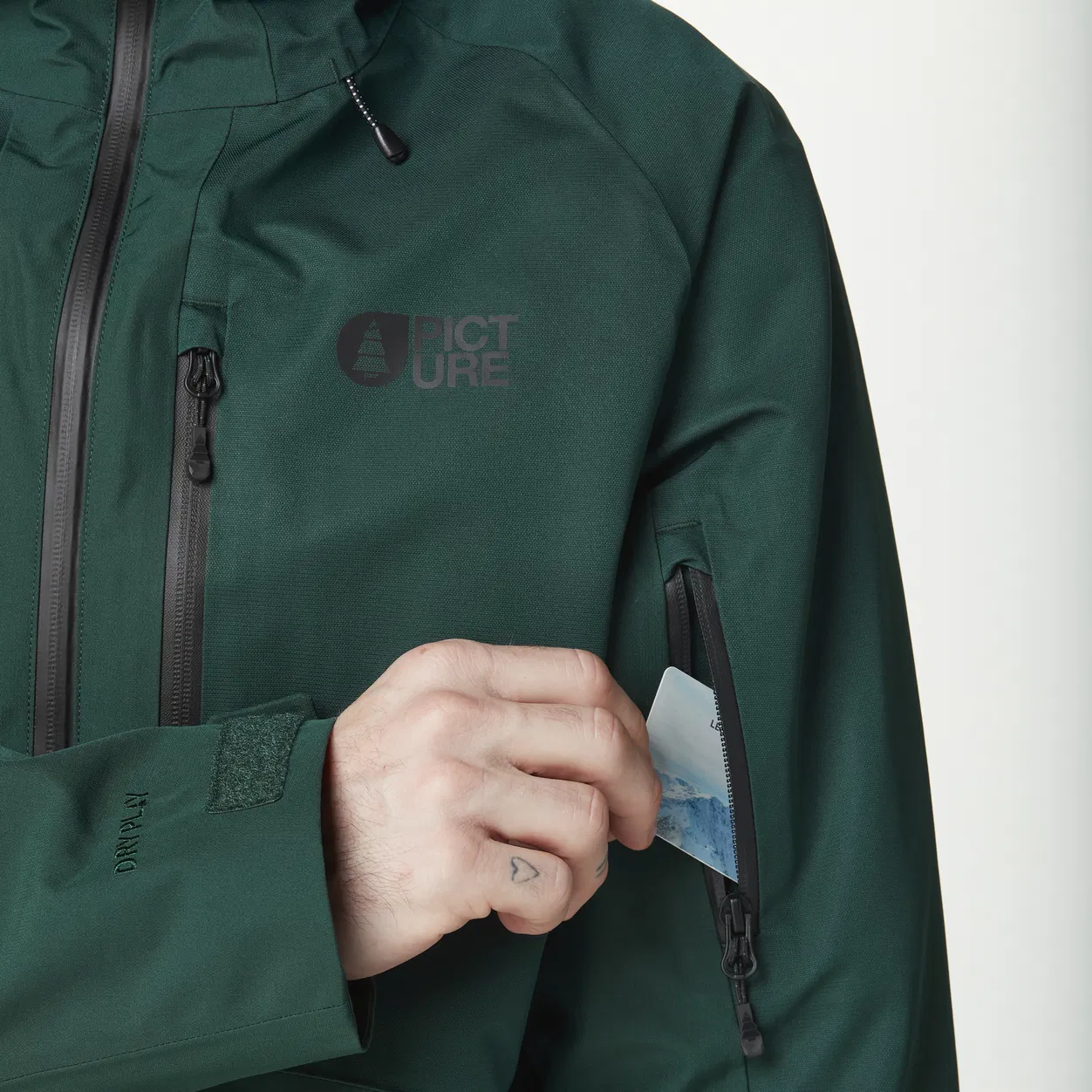 Main d'une personne insérant une petite carte dans la poche du bras d'une veste vert foncé portant le logo « Picture ». La veste est dotée de fermetures à glissière noires et d'une bande velcro sur la manche.