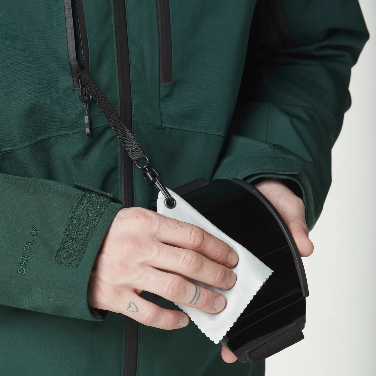 Une personne portant une veste verte nettoie une paire de lunettes de ski avec un chiffon blanc. Le chiffon est attaché à la veste par un cordon. Les mains de la personne ont des tatouages visibles.