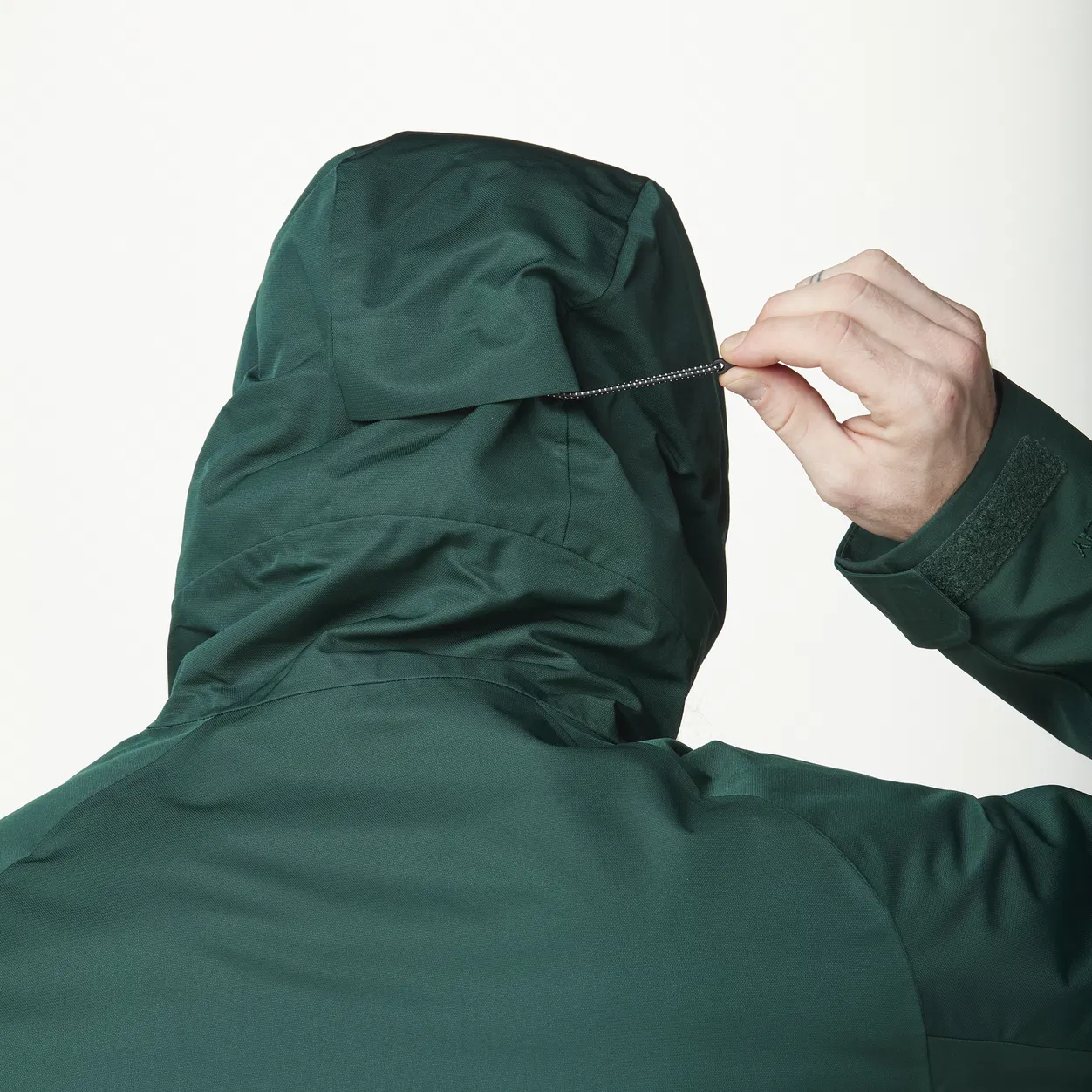 Une personne portant une veste à capuche vert foncé est représentée de dos, ajustant la taille de la capuche à l'aide d'un cordon de serrage. L'arrière-plan est uni et de couleur claire.