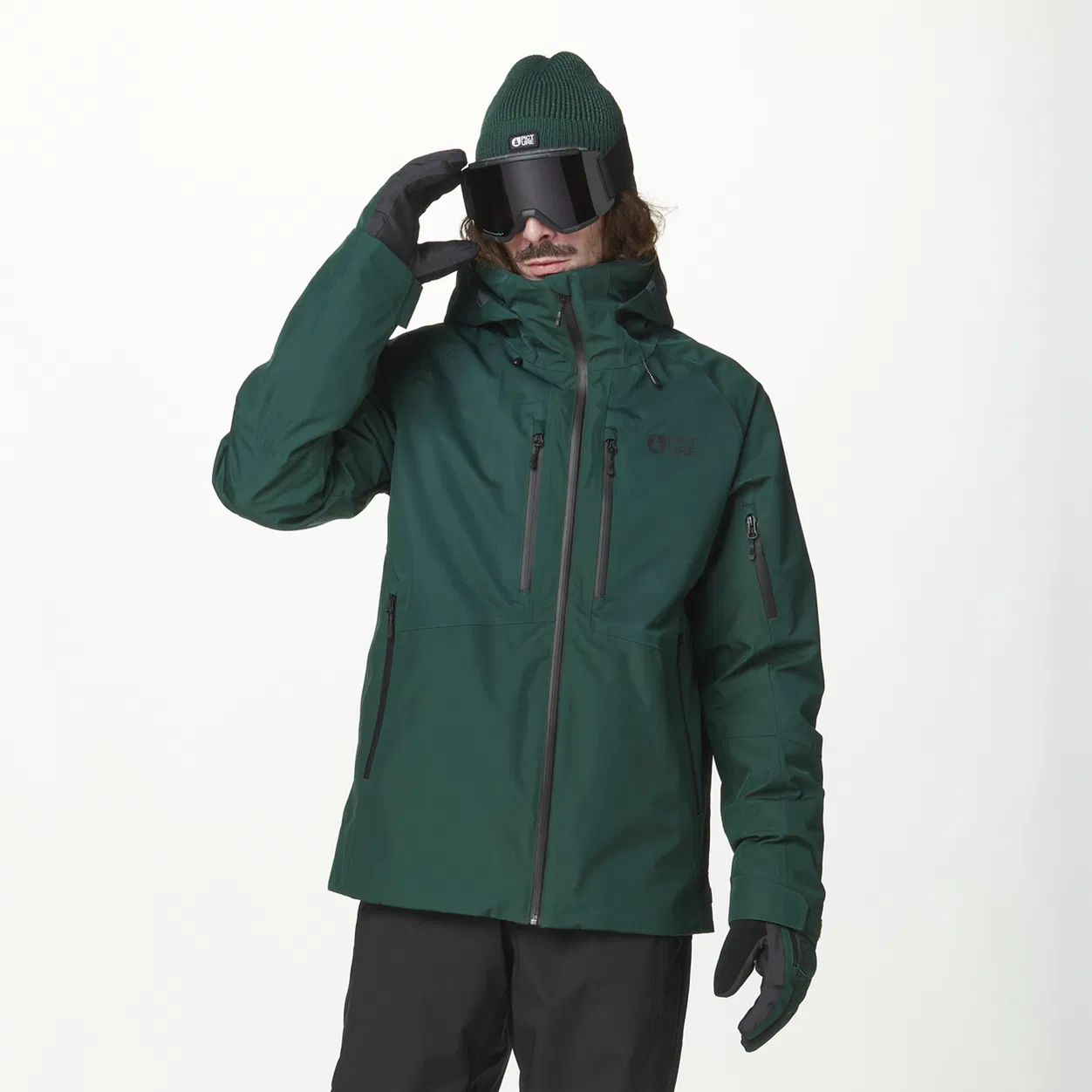 Une personne portant une veste de ski vert foncé, des gants noirs et un bonnet vert porte de grandes lunettes de ski et un masque de ski. Elle ajuste ses lunettes d'une seule main. L'arrière-plan est blanc uni.