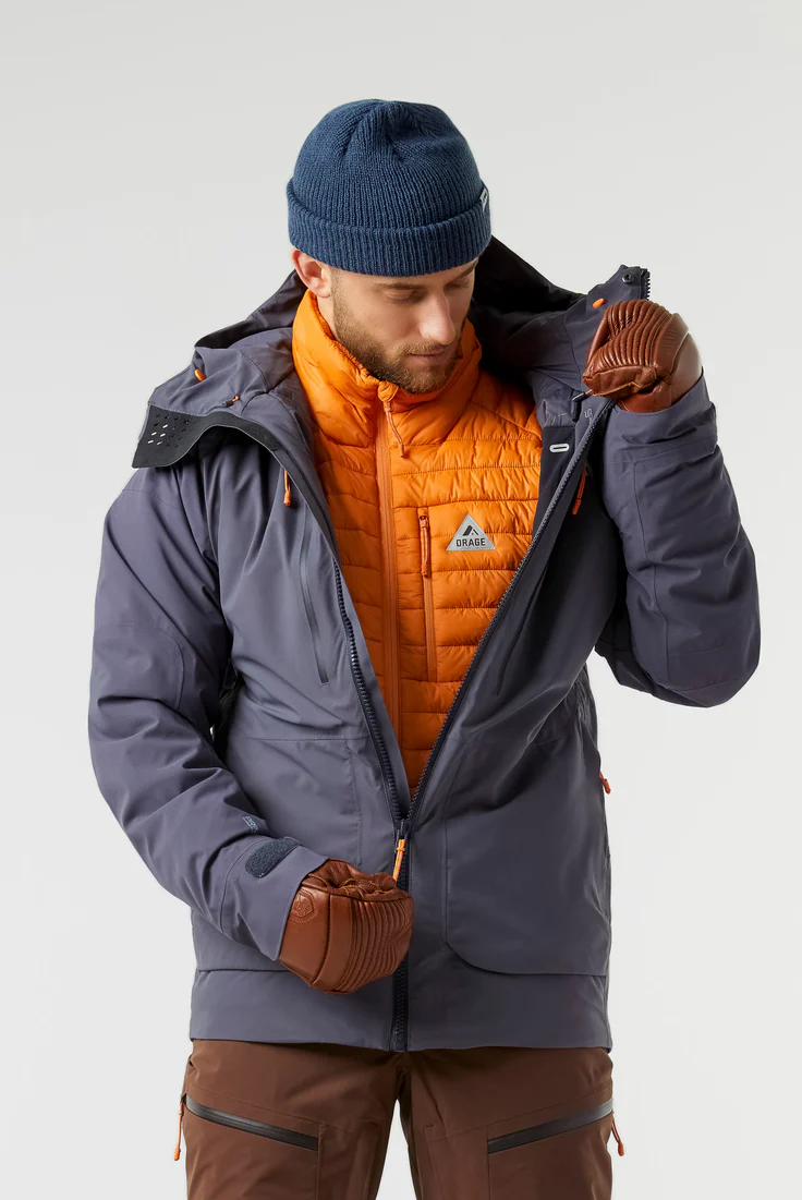 Une personne portant un bonnet bleu, une doudoune orange vif et une veste extérieure grise ajuste sa fermeture éclair. Elle porte également des gants et un pantalon marron. L'arrière-plan est neutre, mettant en valeur sa tenue d'hiver.