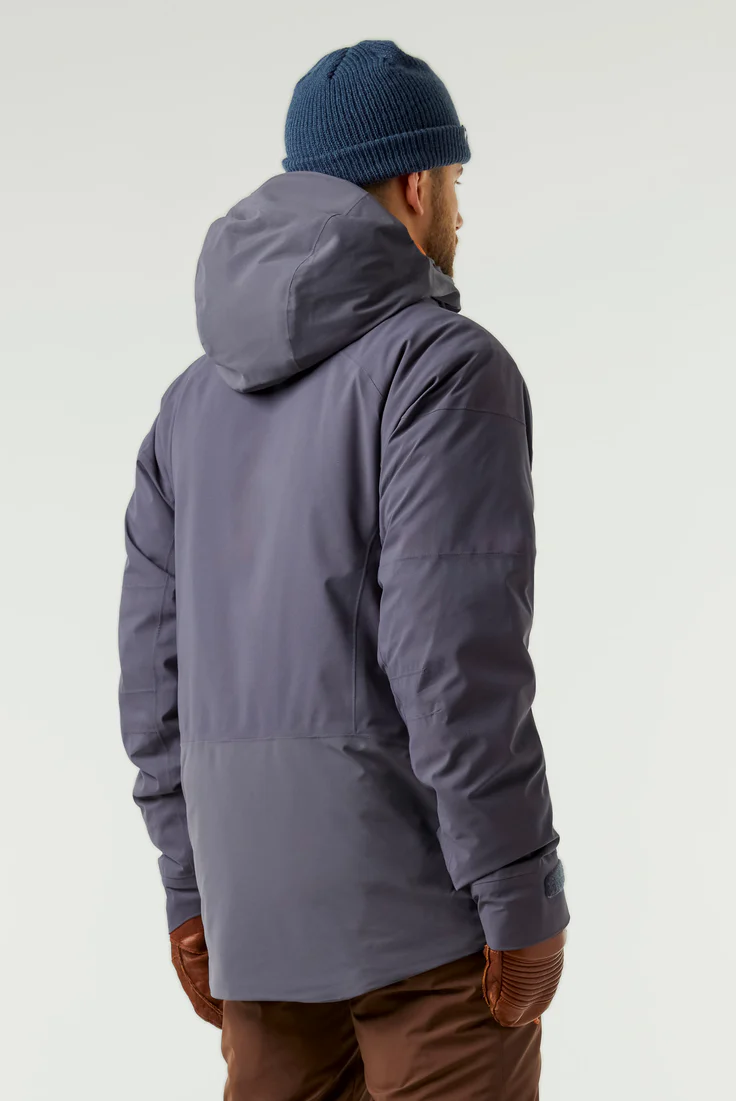 Une personne porte une veste à capuche bleu foncé, un pantalon marron, des gants marron et un bonnet bleu en tricot. Elle tourne le dos à la caméra, debout sur un fond uni et clair.