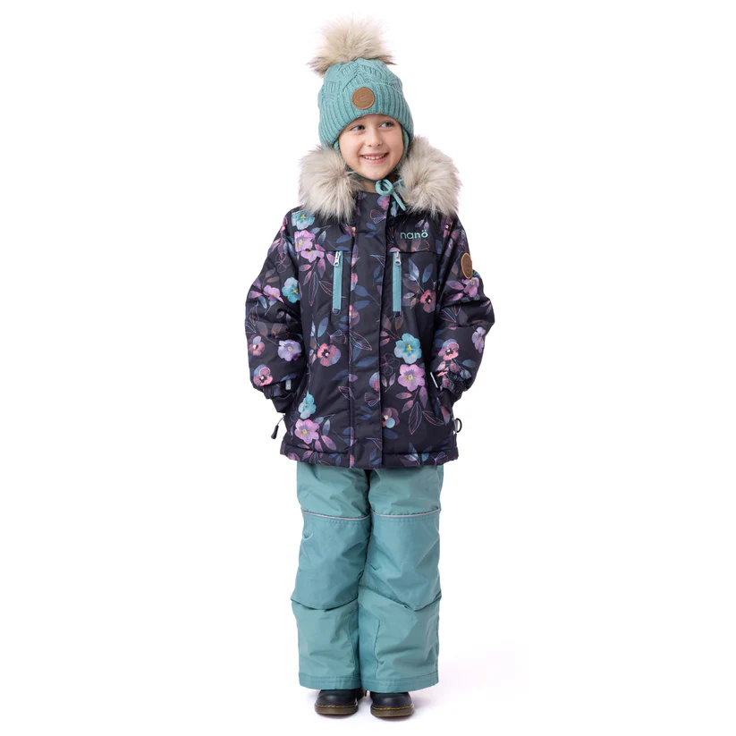 Un enfant sourit tout en portant des vêtements d'hiver. Il porte un manteau à motifs floraux avec une capuche doublée de fourrure, un pantalon de neige bleu sarcelle et un bonnet en tricot bleu sarcelle avec un pompon sur le dessus. Le manteau a plusieurs fermetures éclair et l'enfant a les mains dans les poches.