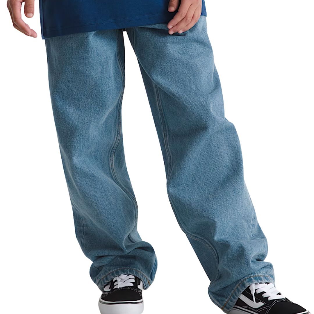 Vêtu d'un pantalon en jean ample B CHECK-5 de couleur bleu clair, cet individu se tient debout, les jambes écartées, et porte des baskets noires et blanches. L'image se concentre sur les jambes et une partie d'une main ; le haut du corps n'est pas visible.