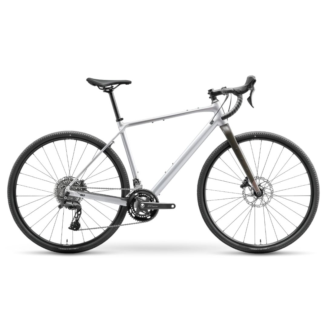 Le VÉLO DE GRAVEL NORCO SEARCH A CUES est un vélo de gravier argenté avec un guidon tombant, des freins à disque et une selle noire, présenté de côté sur un fond blanc.