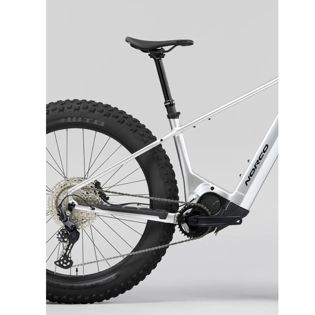 Vue latérale d'un vélo électrique Norco argenté montrant la roue arrière, le siège et le moteur à entraînement central. Il est doté de pneus épais avec une chaîne et un dérailleur visibles. Le vélo est placé sur un fond gris uni.
