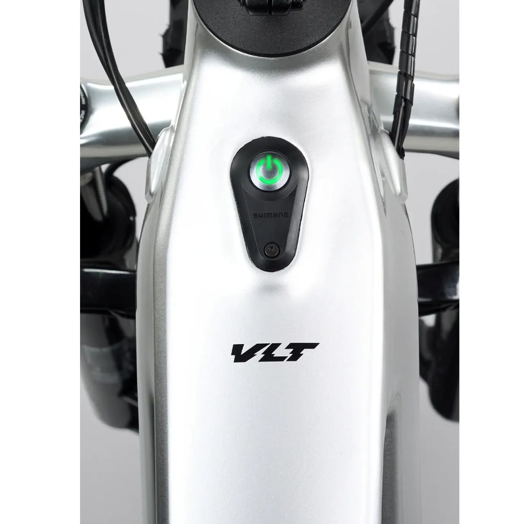 Gros plan sur un cadre de vélo argenté avec un bouton d'alimentation noir et une lumière verte au-dessus du logo « VLT ». Le guidon est partiellement visible sur les bords de l'image.