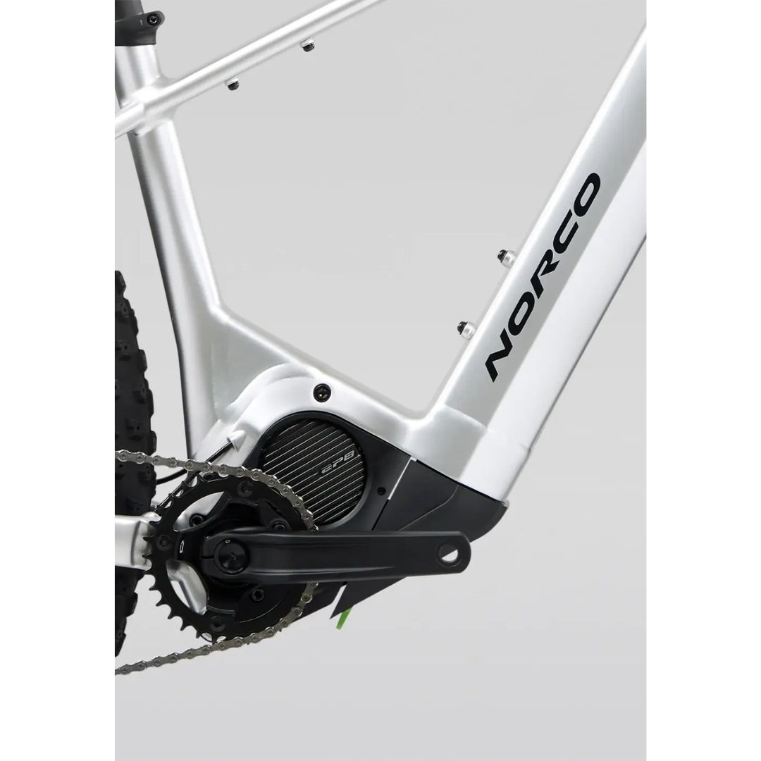 Gros plan de la transmission et du moteur d'un vélo électrique Norco argenté. L'image montre la manivelle, la chaîne et le pneu arrière partiellement visibles sur un fond uni.