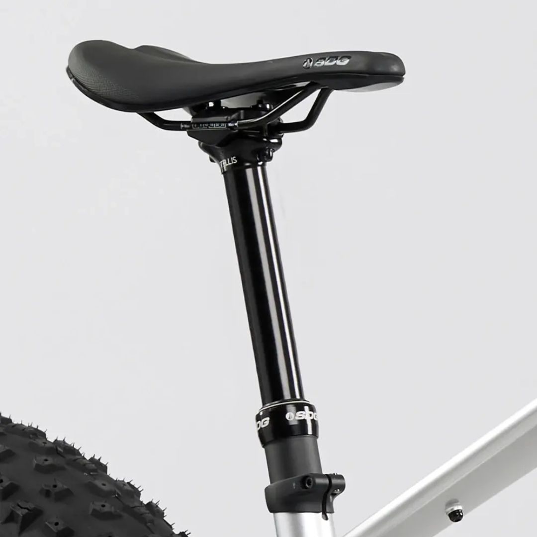 Gros plan d'une selle de vélo avec une tige de selle noire fixée sur un cadre argenté. Le pneu est visible en bas à gauche, avec un motif de bande de roulement épais. L'arrière-plan est d'une couleur unie, gris clair.