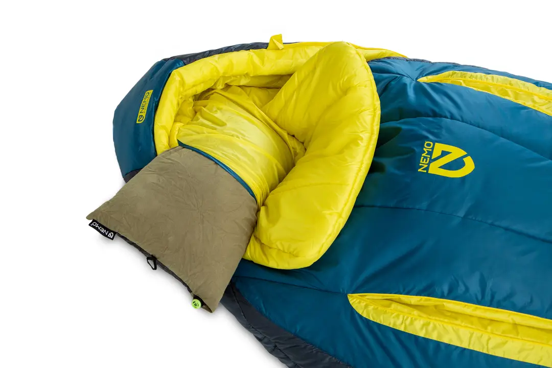 Sac de couchage bleu et jaune avec oreiller intégré et capuche marron amovible, présenté partiellement ouvert pour révéler son intérieur doux et isolant. Le logo NEMO est visible à l'extérieur.