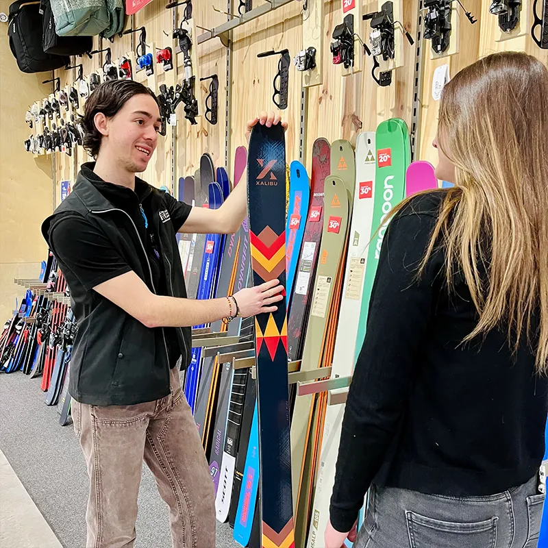 Un employé de magasin montre une paire de skis à une femme dans une boutique de sport, avec différents skis colorés exposés verticalement sur le mur derrière eux.