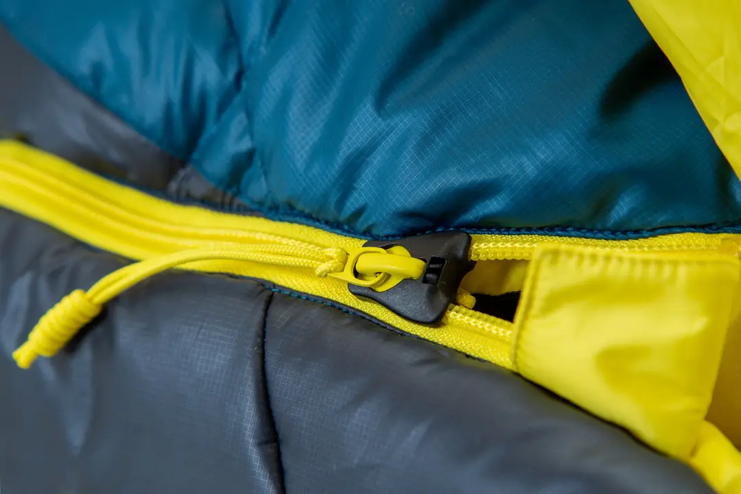 Gros plan d'une fermeture éclair jaune sur un sac de couchage avec des panneaux en tissu bleu sarcelle, gris et jaune.
