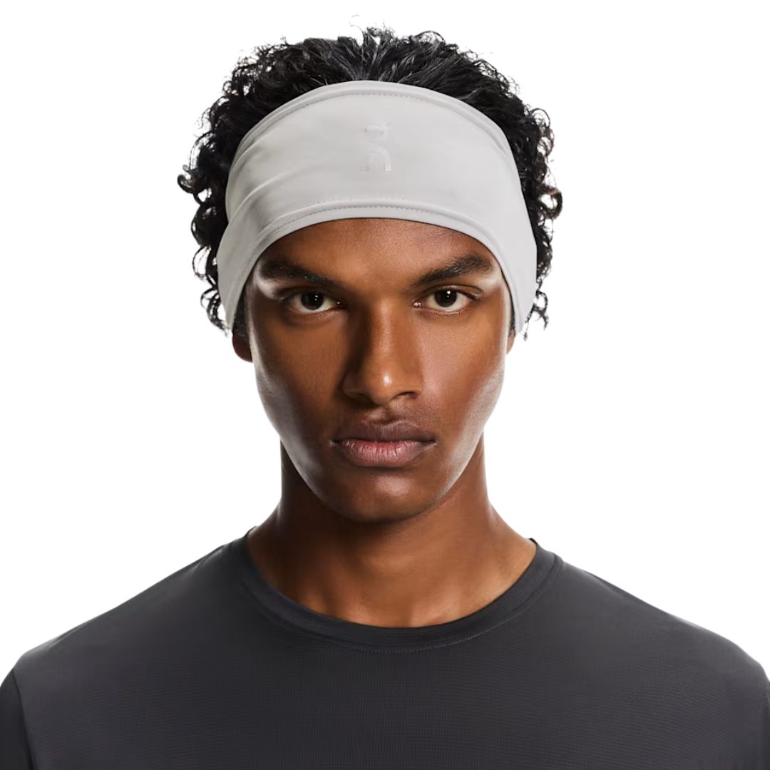 Une personne aux cheveux bouclés porte le bandeau CORE HEADBAND 1 U de couleur gris clair et l'associe à un t-shirt gris foncé ; elle regarde l'appareil photo devant un fond blanc uni.