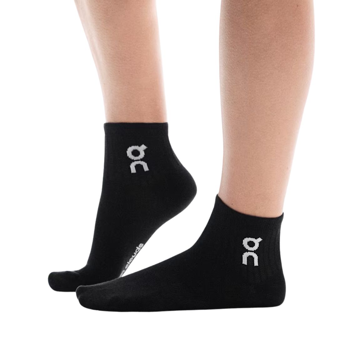 Une personne portant des socquettes noires ornées d'un logo blanc sur le côté, debout sur la pointe des pieds, les deux pieds bien visibles sur un fond blanc.