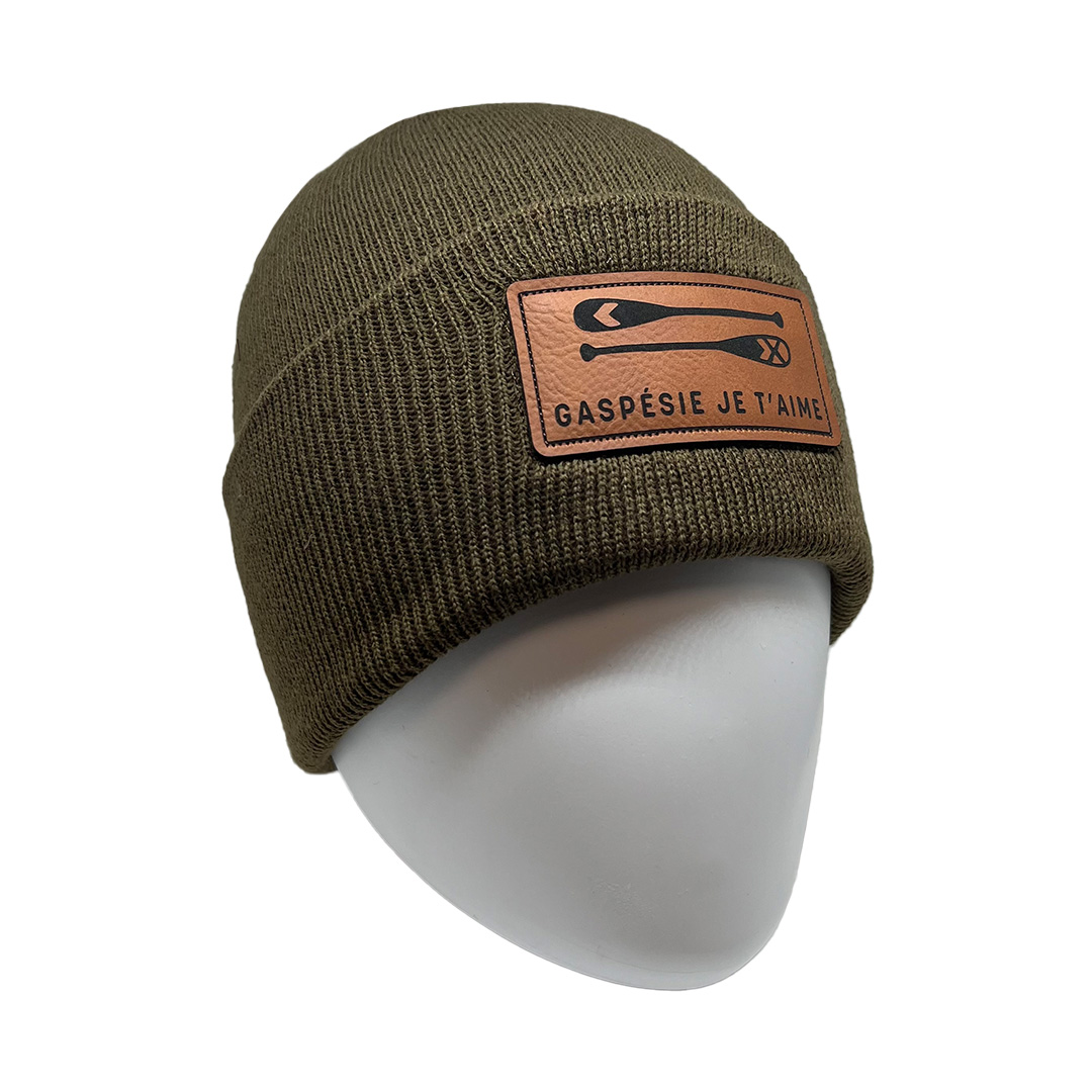 Bonnet en tricot vert olive avec un patch marron représentant des pagaies de canot croisées et les mots « Gaspésie Je T'aime ». Le bonnet est présenté sur une tête de mannequin blanche sur fond uni.