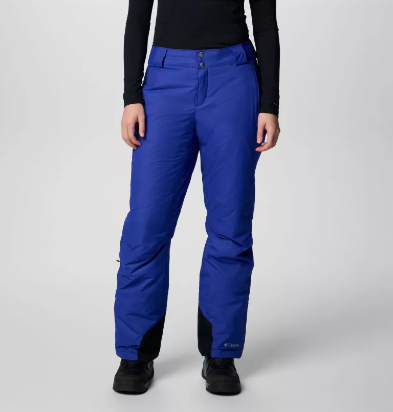Une personne portant un haut noir à manches longues associé à un pantalon imperméable bleu vif et à des chaussures noires se tient debout sur un fond blanc uni. Le pantalon a des revers noirs aux chevilles et de multiples poches.