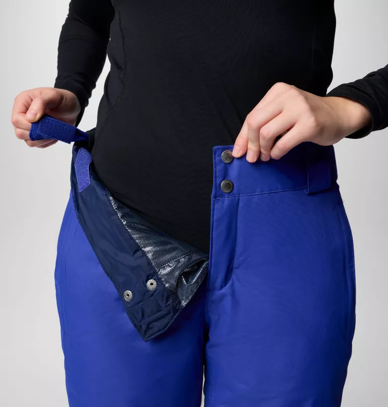 Une personne portant un haut noir à manches longues montre comment ajuster un pantalon bleu avec des attaches velcro et boutonnées.