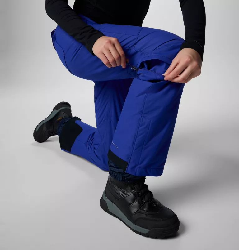 Une personne s'agenouille tout en ajustant la fermeture éclair de la partie inférieure d'un pantalon convertible bleu cobalt. Elle porte des bottes de randonnée noires et un haut noir à manches longues. Le fond de la photo est blanc uni.