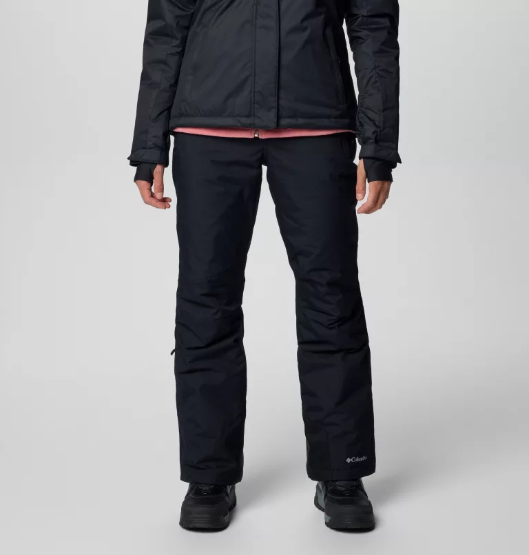 Une personne portant une veste noire et un pantalon noir se tient debout sur un fond blanc uni. Ses mains sont visibles et elle porte des chaussures noires. Les vêtements semblent adaptés au temps froid.