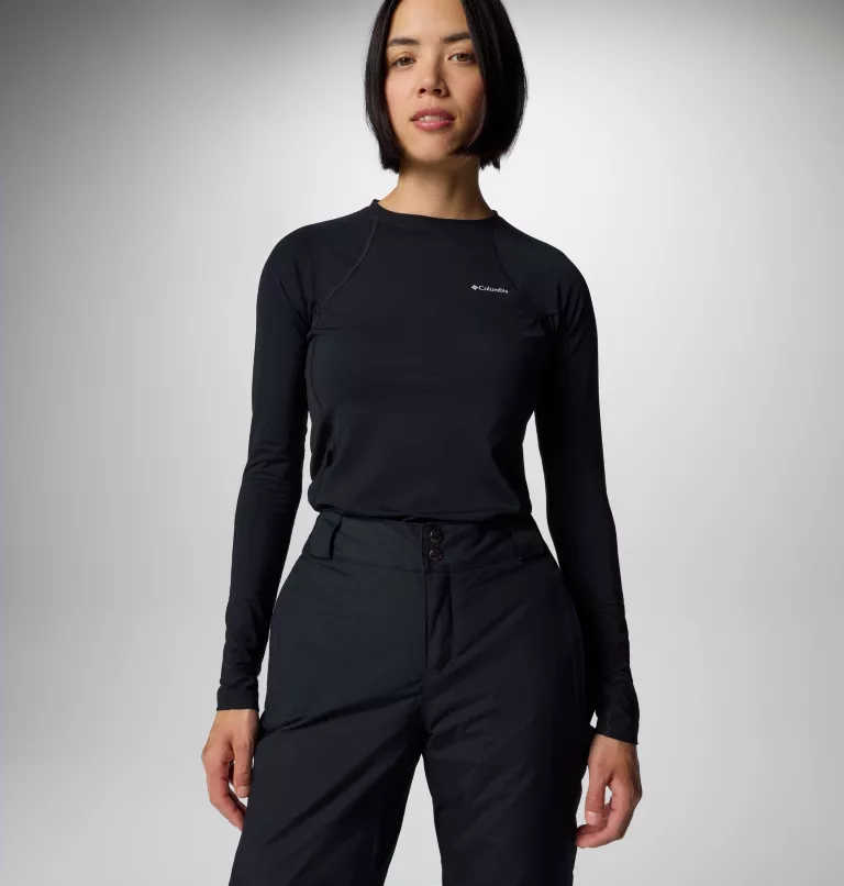 Une personne portant un haut à manches longues noir et un pantalon noir se tient debout avec assurance sur un fond clair uni. La tenue comprend un haut de marque avec un petit logo sur la poitrine.