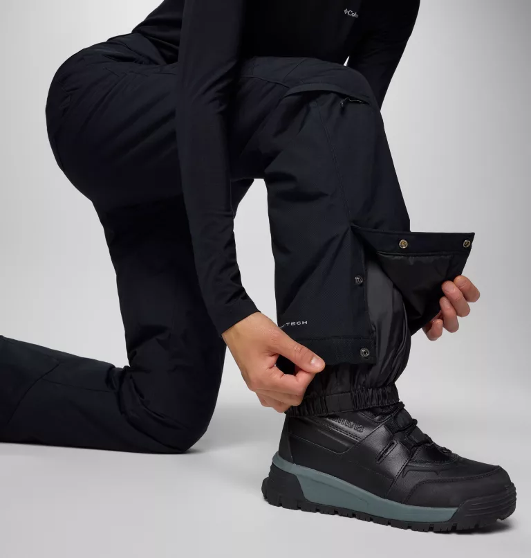 Une personne s'agenouille tout en ajustant le revers de son pantalon noir, révélant une botte noire et grise montant jusqu'à la cheville. Le pantalon comporte des boutons visibles et une fermeture éclair, et le fond est blanc uni.