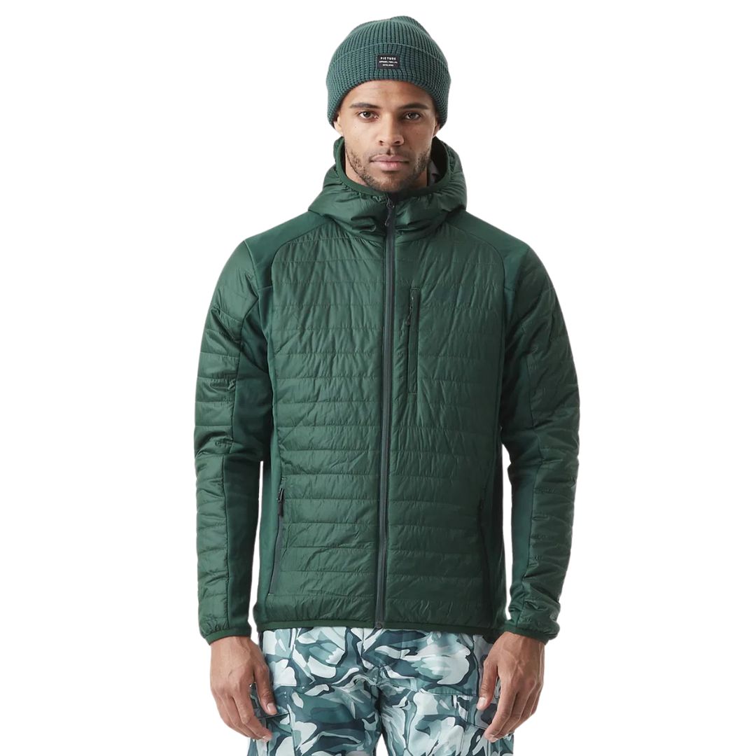 Une personne porte une veste matelassée verte avec une capuche et un bonnet en tricot vert assorti. Elle porte un pantalon à motif camouflage bleu et blanc. Le fond est blanc uni, ce qui met en valeur la tenue d'extérieur.