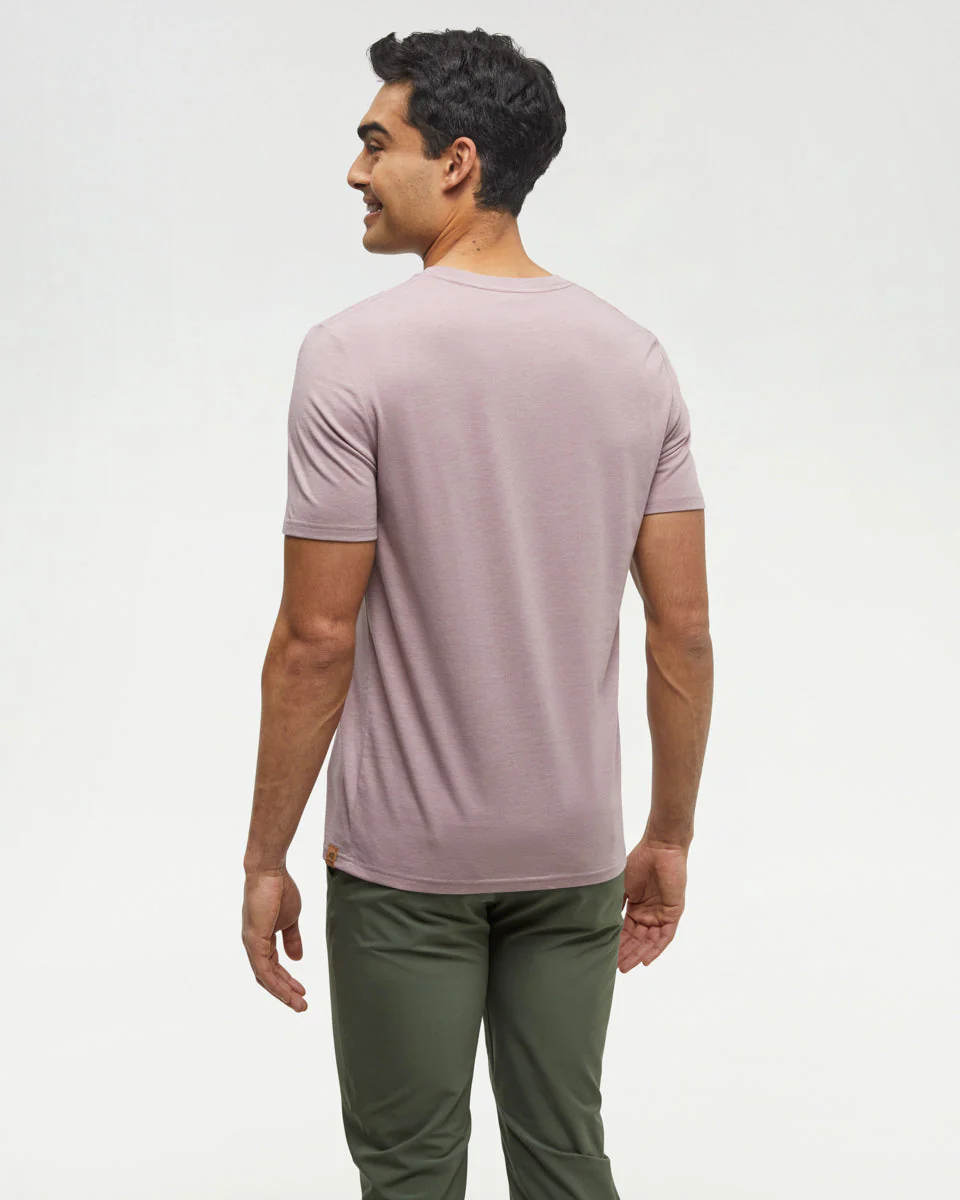 Un homme aux cheveux noirs courts est debout et tourné vers l'extérieur, vêtu d'un t-shirt à manches courtes mauve clair et d'un pantalon vert olive, sur un fond clair uni.