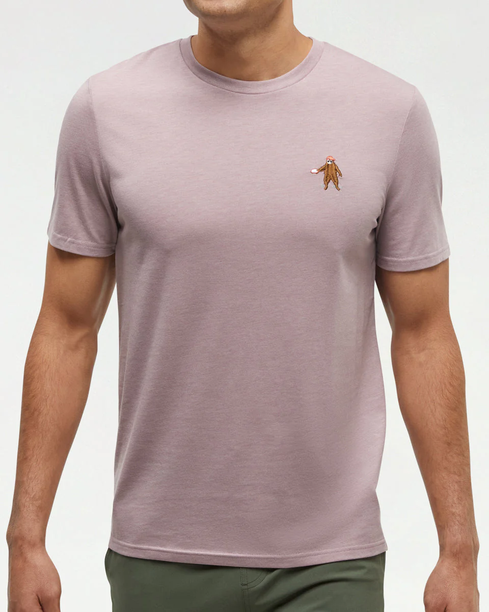 Un homme portant un t-shirt à manches courtes mauve clair avec une petite figure brodée sur la poitrine gauche, associé à un pantalon vert foncé, debout sur un fond clair uni.