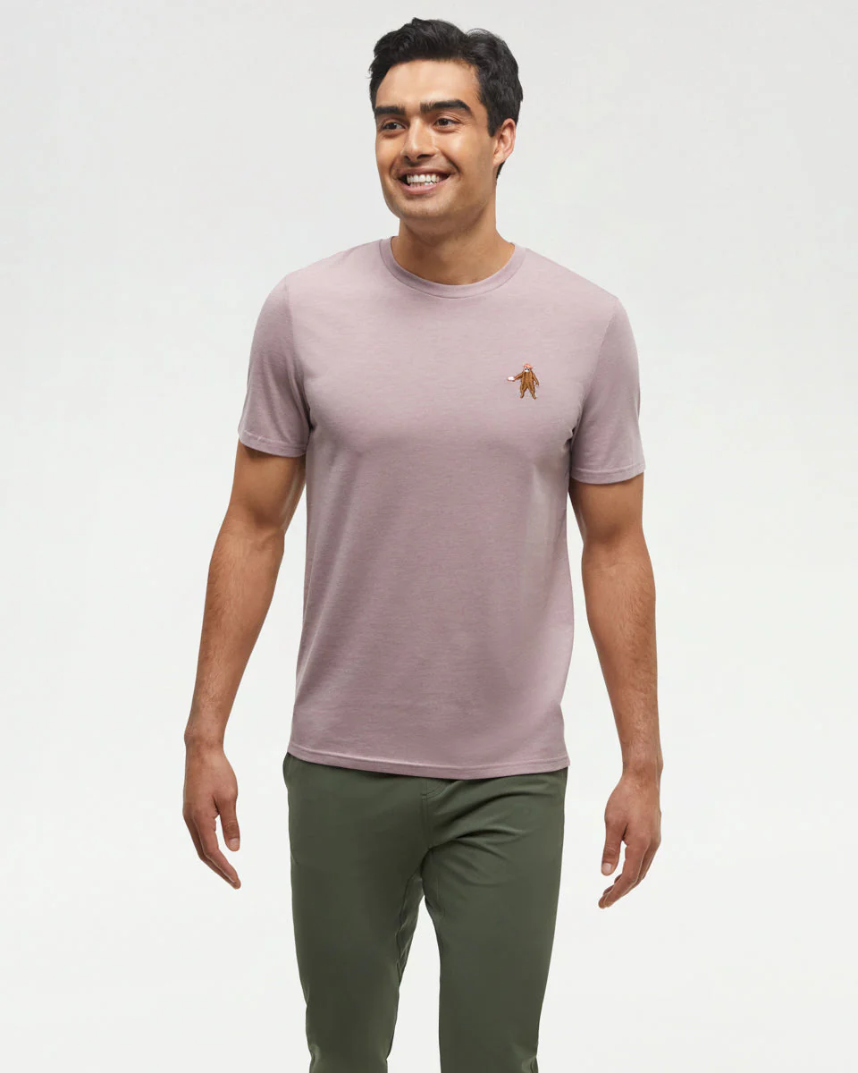 Un homme aux cheveux noirs courts sourit et se tient sur un fond clair, portant un t-shirt mauve clair avec un petit logo brodé sur la poitrine et un pantalon vert olive.