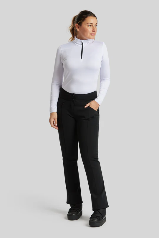 Une femme se tient debout sur un fond gris uni. Elle porte un haut blanc à manches longues et à col montant avec une fermeture éclair noire et un pantalon noir. Ses cheveux sont tirés en arrière en queue de cheval et elle regarde légèrement vers la gauche avec ses mains dans ses poches.