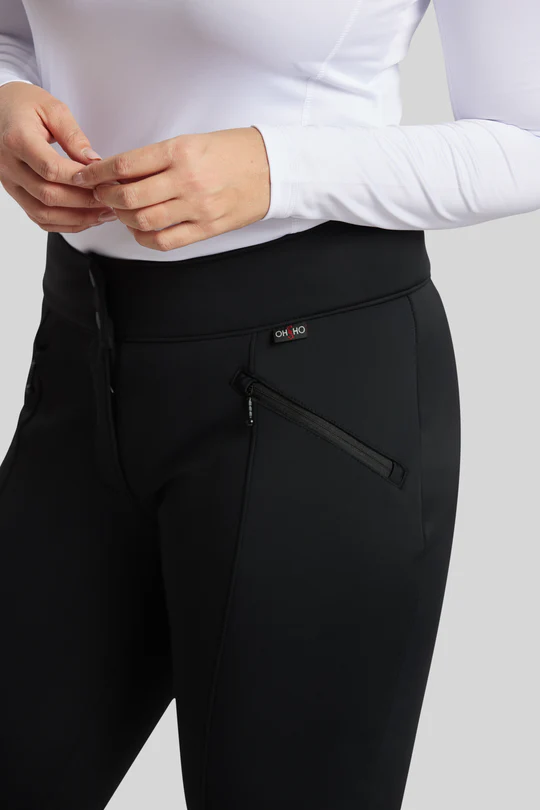 Une personne porte un pantalon noir à taille haute avec des poches zippées et un haut blanc à manches longues. Seuls son torse et une partie de ses bras sont visibles. Le pantalon comporte un petit logo « OKHO » sur la ceinture. Le fond est gris uni.