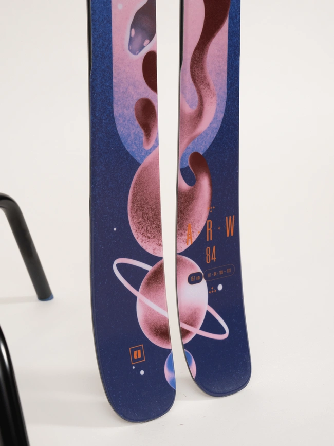Gros plan de deux skis au design à thème cosmique avec des tourbillons roses et violets, des planètes et des étoiles. Les skis portent le texte « A R W 84 ».