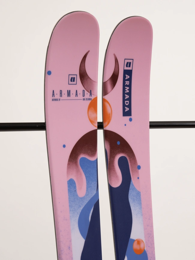 Une paire de skis roses avec des motifs abstraits en bleu, marron et orange. La marque « Armada » est imprimée sur chaque ski, ainsi que des graphiques circulaires et des motifs en forme de vagues. Les skis sont positionnés à la verticale sur un fond uni.