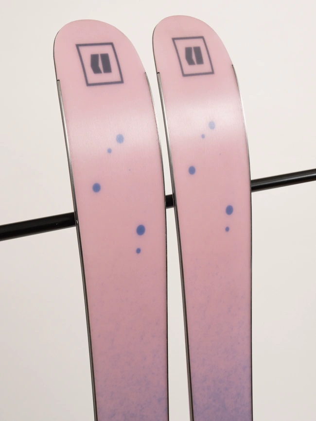 Une paire de skis roses à pois bleus appuyée contre un bâton noir. Les pointes des skis portent un logo géométrique près du haut. Le fond est neutre et uni.