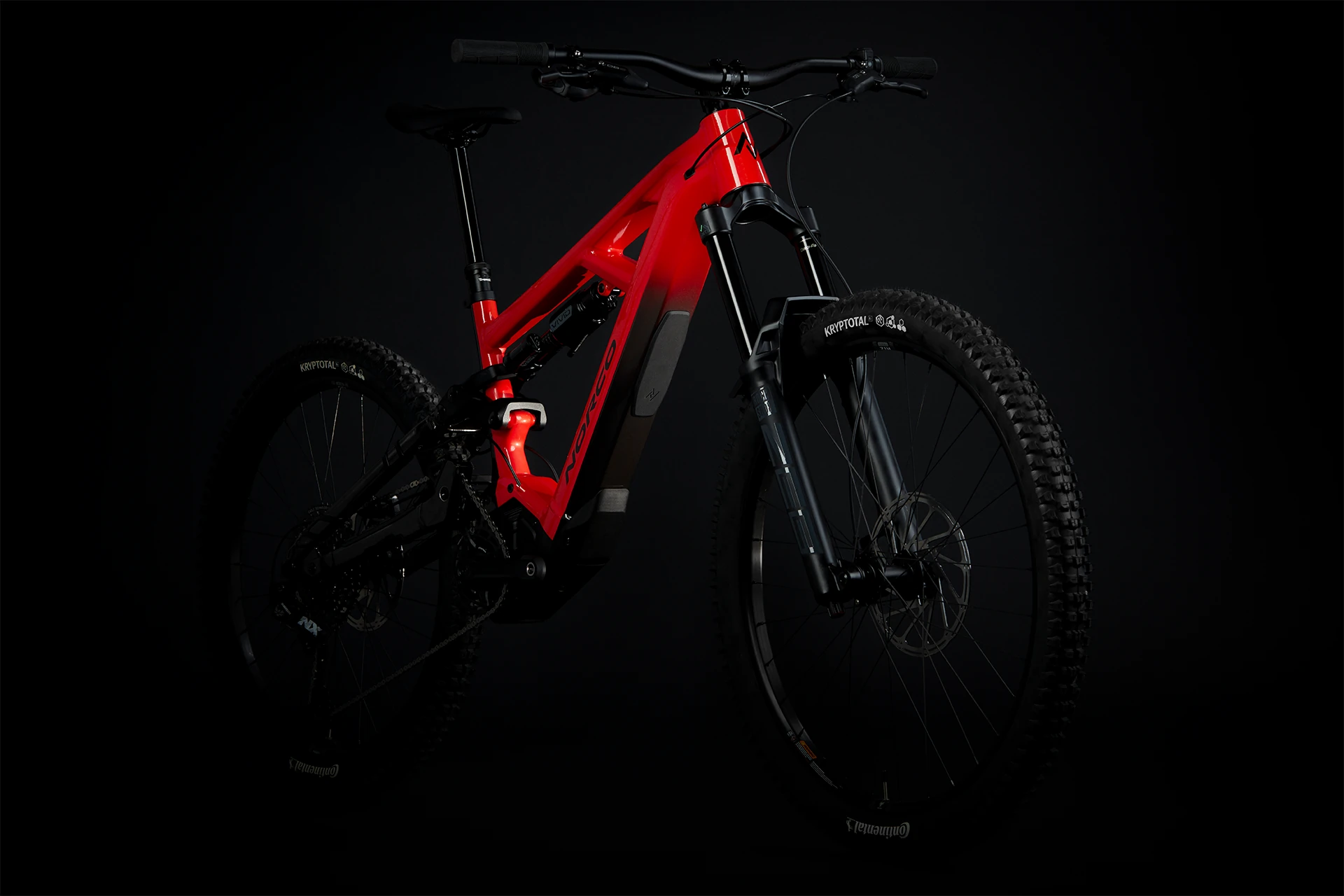Un VTT rouge et noir, élégant et moderne, avec des pneus épais et une suspension intégrale, photographié sur un fond sombre. Le vélo est incliné pour révéler les détails du cadre et de la fourche avant.
